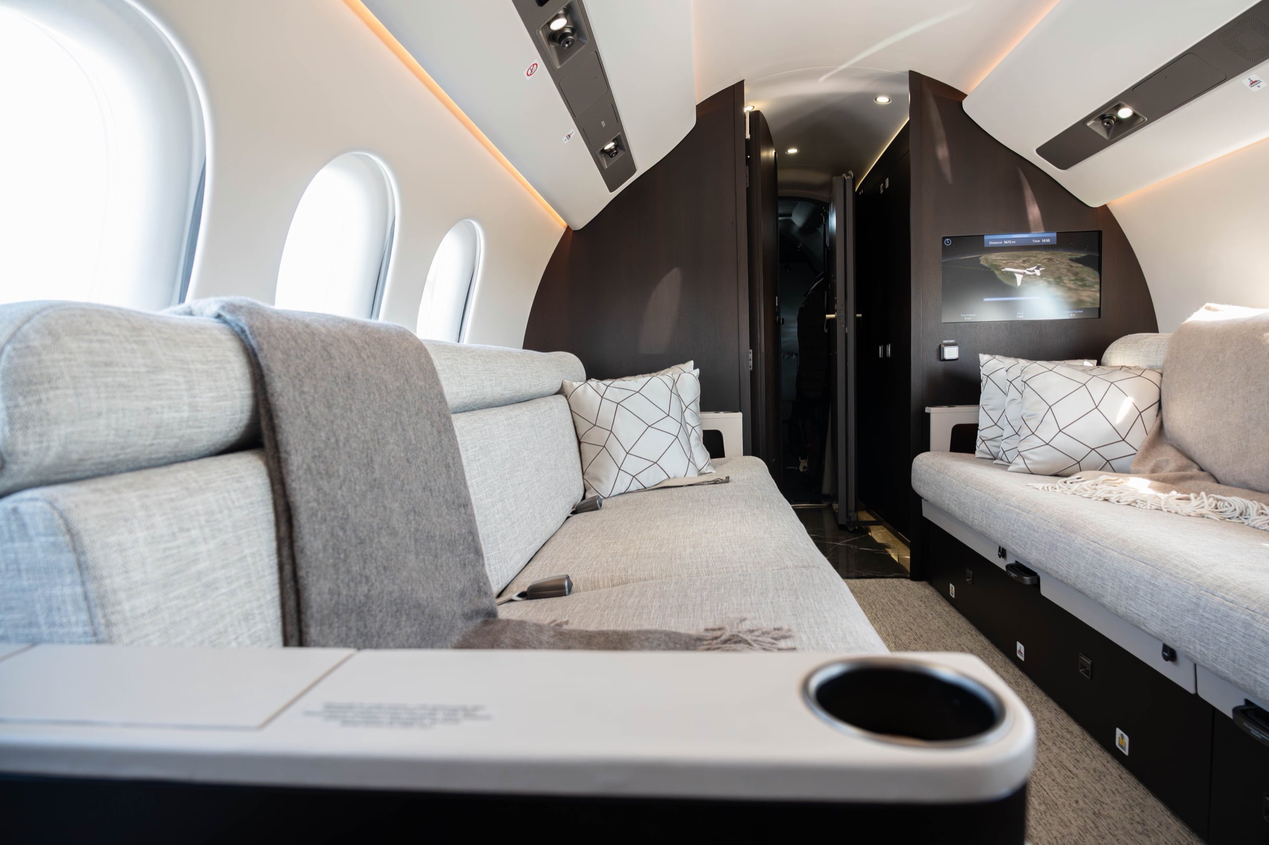 A private jet cabin divan dressed in grey wool, flight display glowing beyond