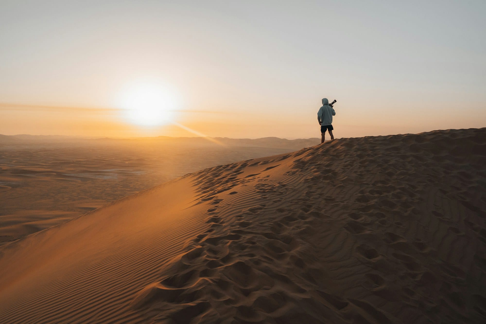 A lone figure stands on a rippled dune ridge as the sun sets over the Dubai desert