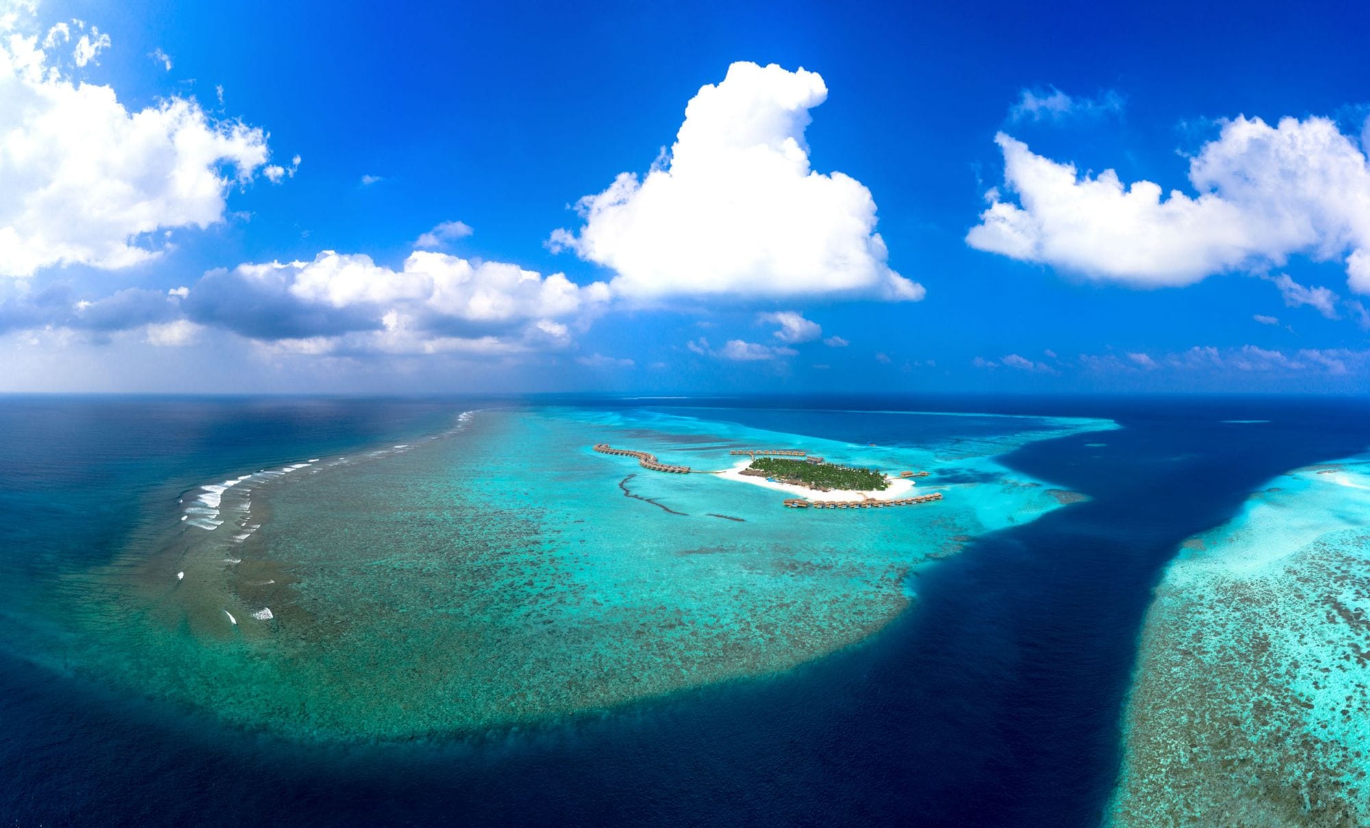 A private island resort ringed by coral reef and glowing lagoon water under towering clouds
