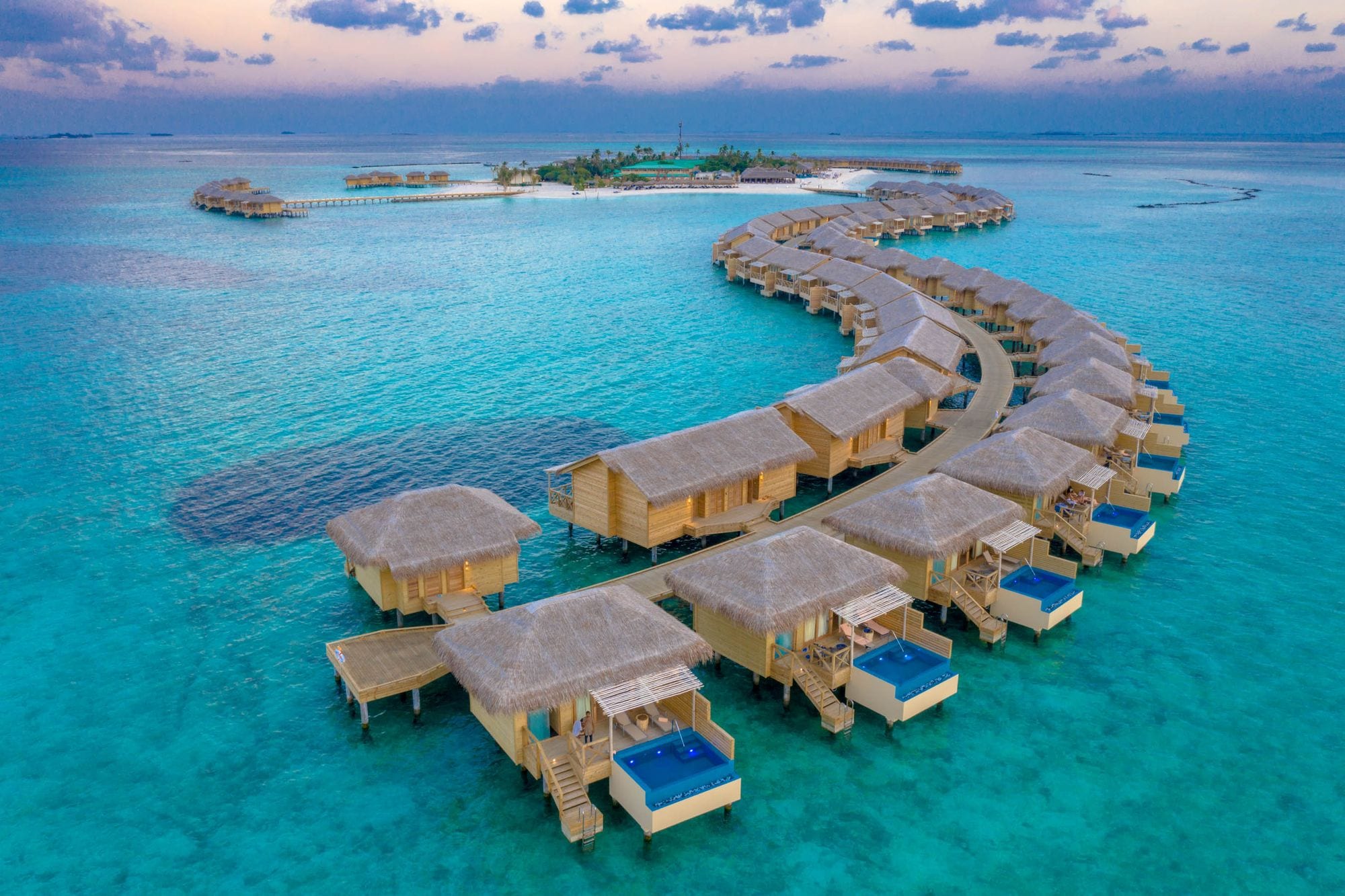 Thatched overwater villas with private pools curve through a turquoise lagoon at dusk