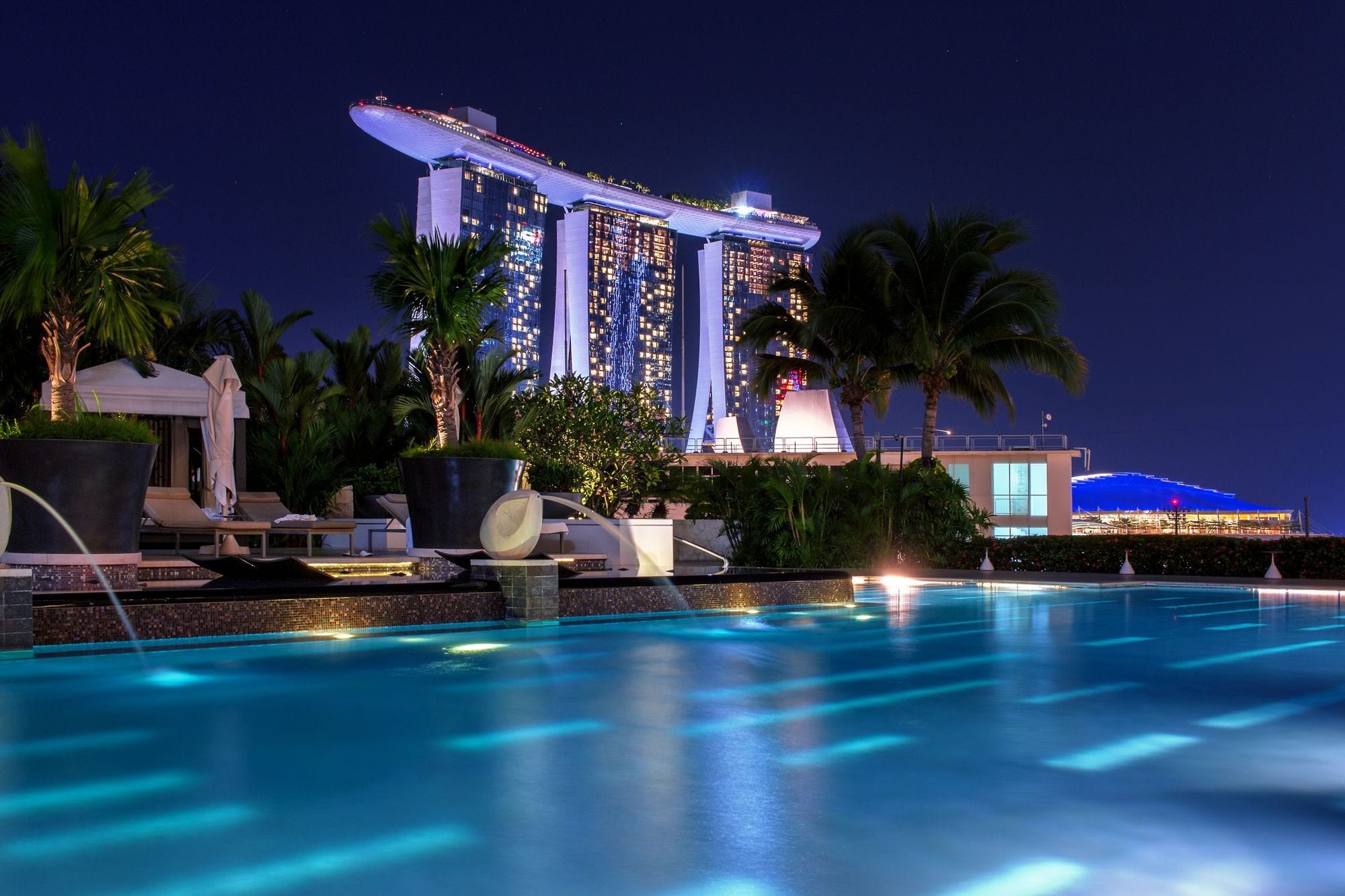 A palm-fringed hotel pool shimmers at night beneath the illuminated towers of Marina Bay Sands