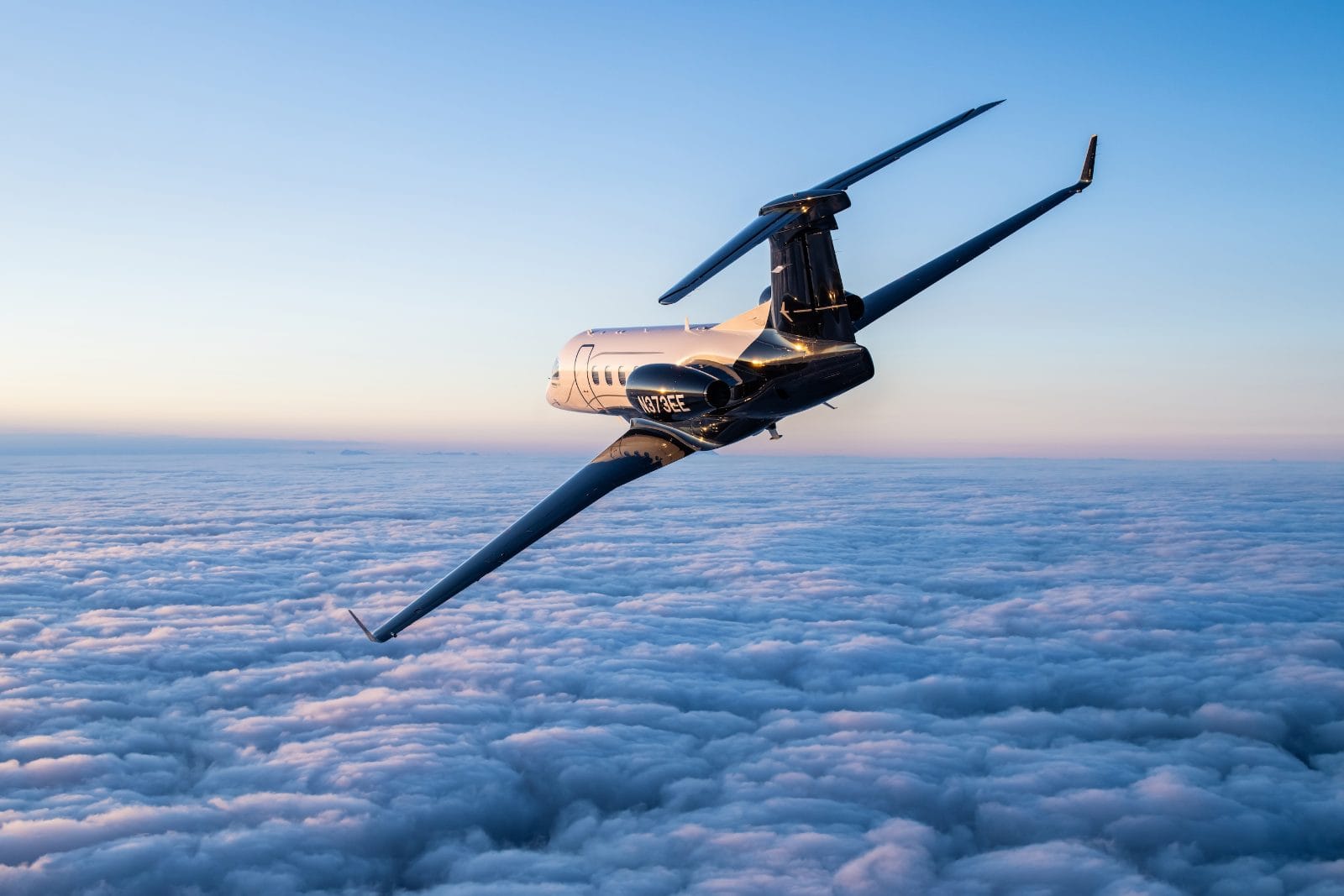 The Embraer Phenom 300EV banking over a sea of cloud at dawn, its dark fuselage catching the first light