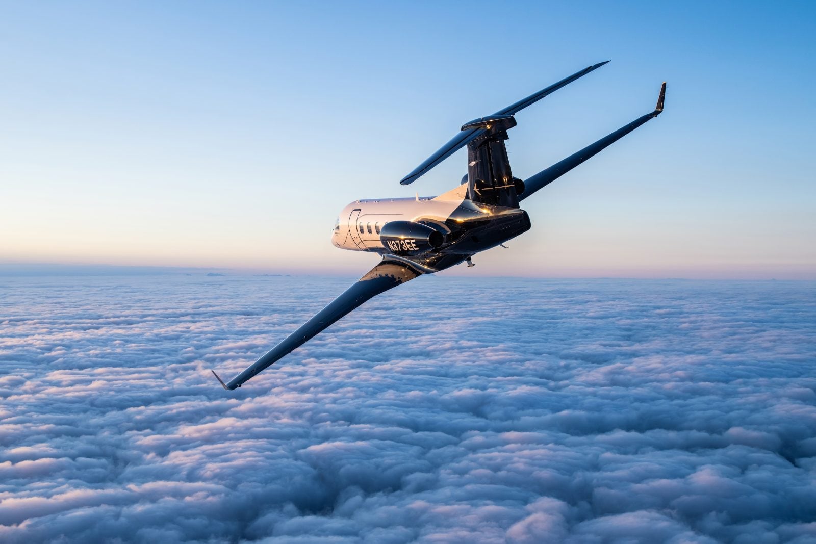 The Embraer Phenom 300EV banking over a sea of cloud at dawn, its dark fuselage catching the first light